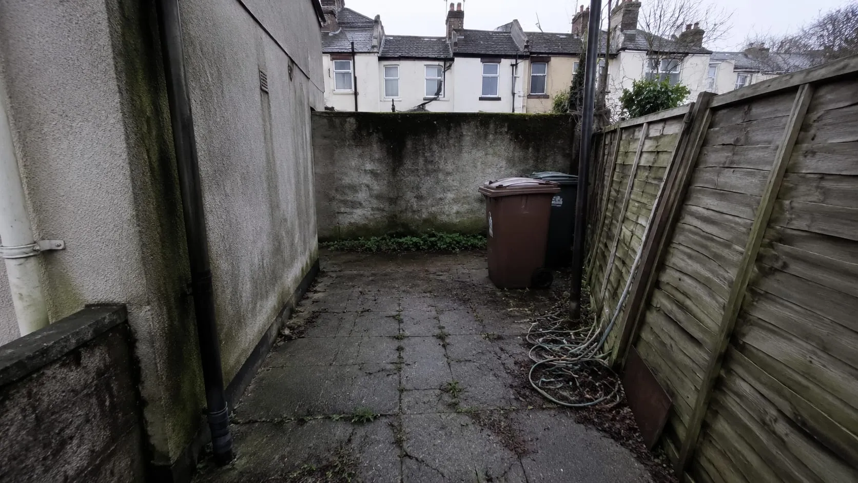 A neglected narrow side-return yard before work: cracked concrete with weeds through the joints, wheelie bins and a leaning rotten fence panel.