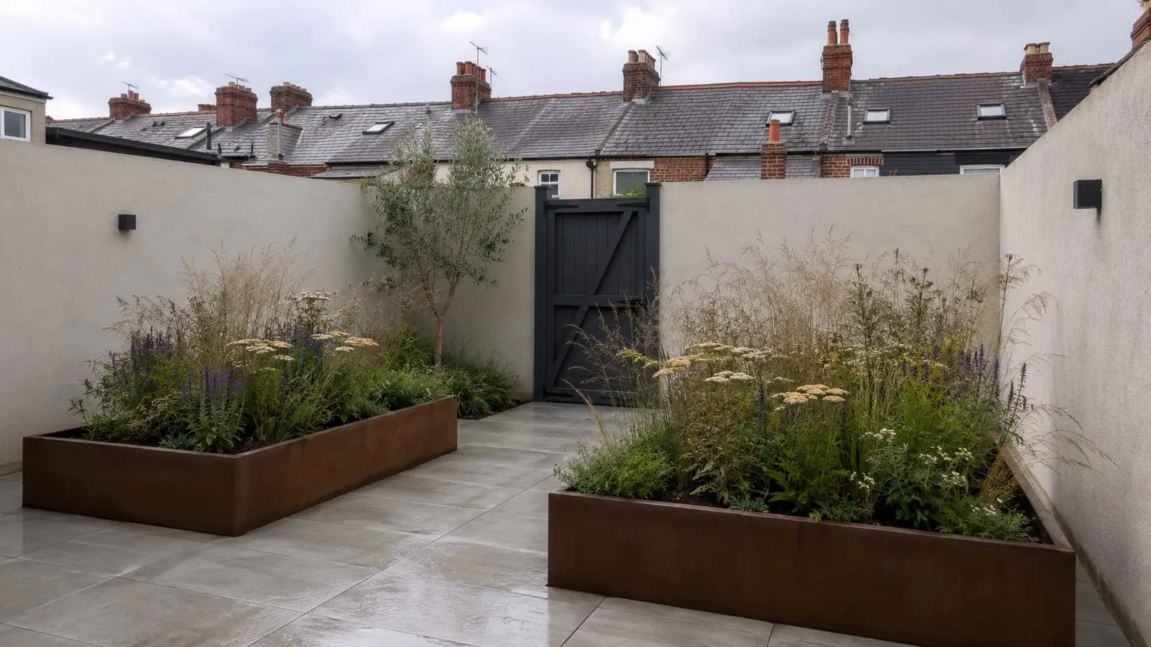 An enclosed courtyard garden paved in pale porcelain, with two weathered corten steel planters full of grasses and salvia, terraced rooftops beyond the wall.